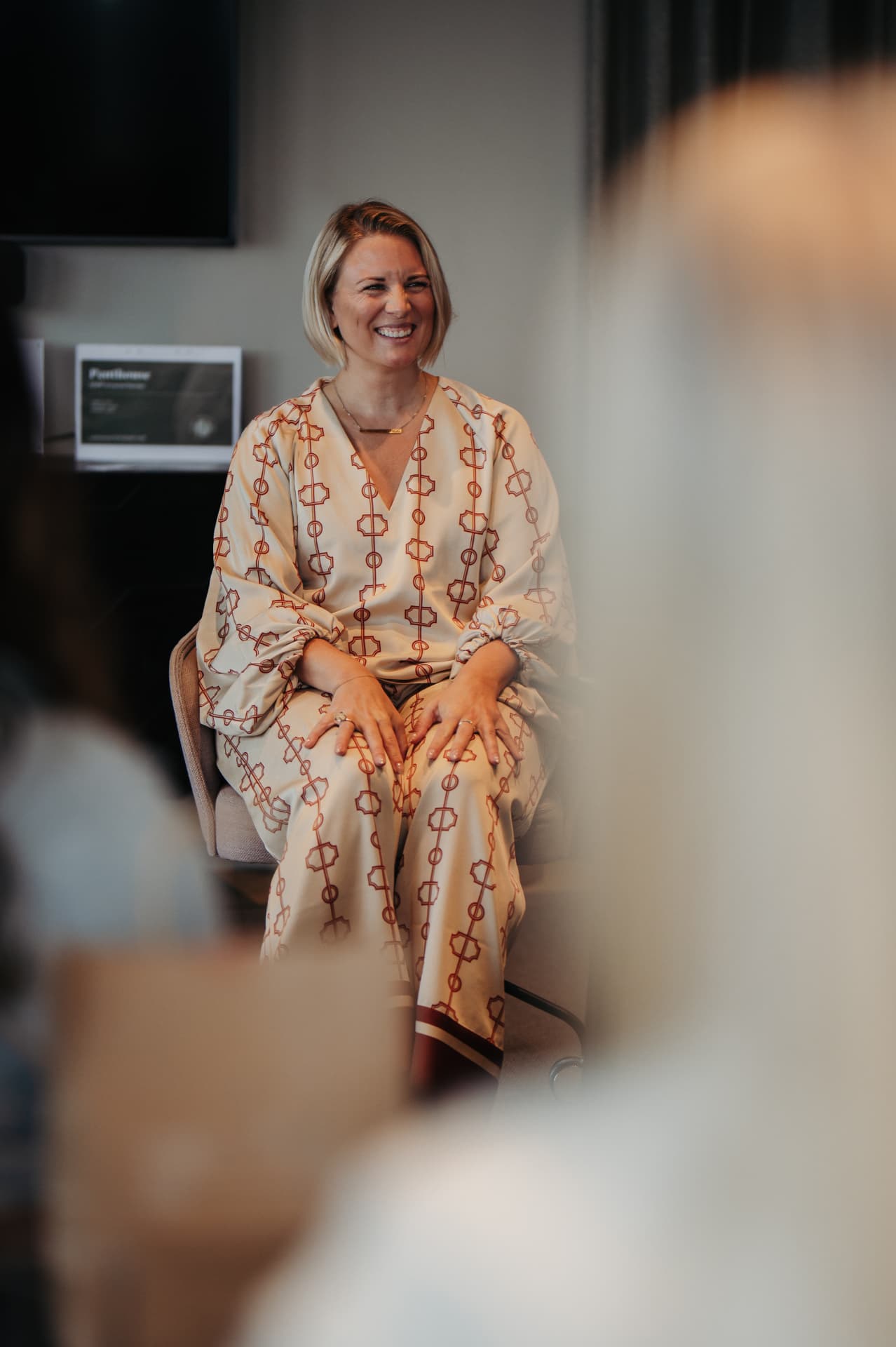 Amanda Randau sitter i en stol i ett mönstrat ljusbeige set och ler under ett samtal, fotograferad över axeln på åhörare i förgrunden